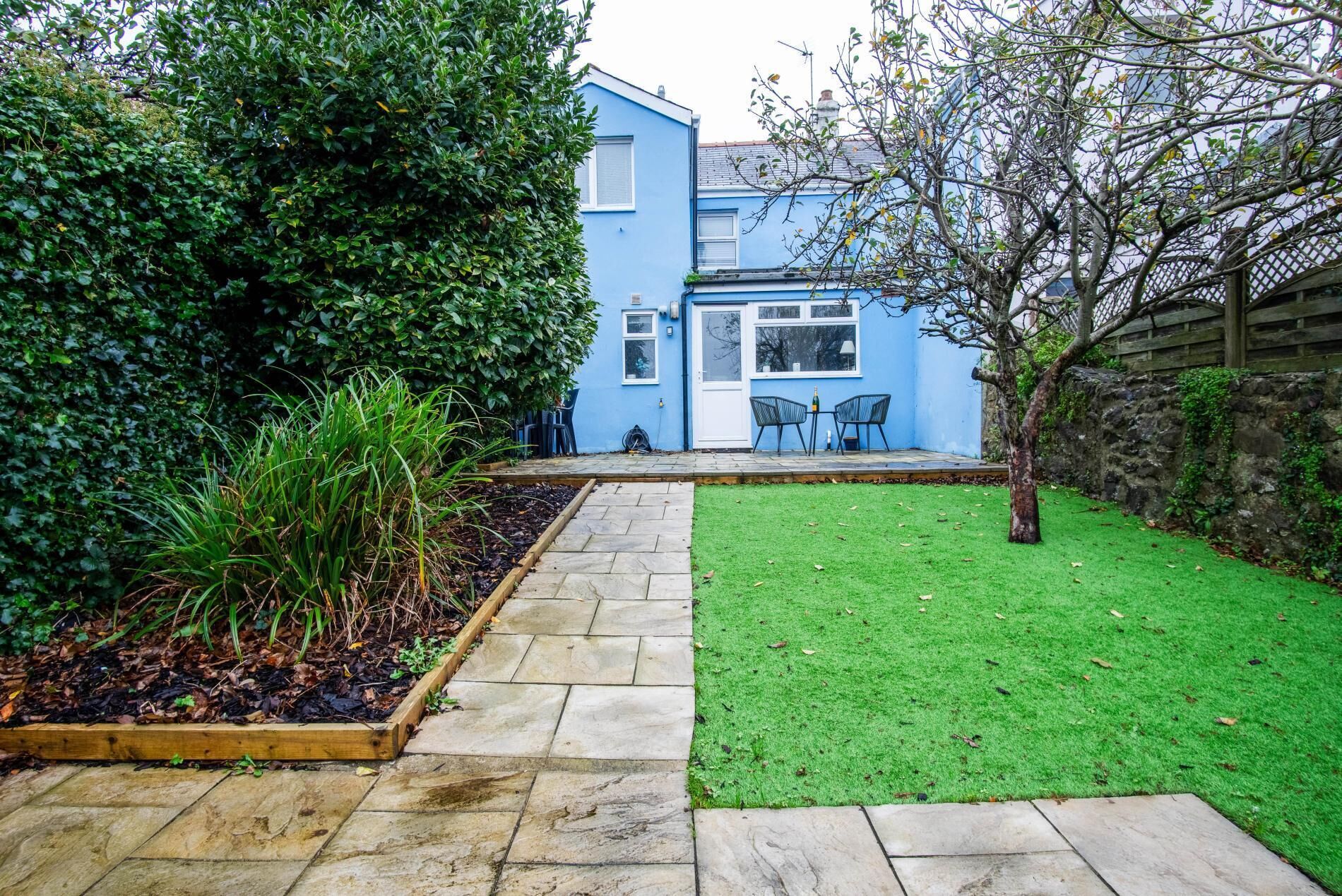 A blue house with a paved pathway leading to a grassy area and a small patio with chairs.