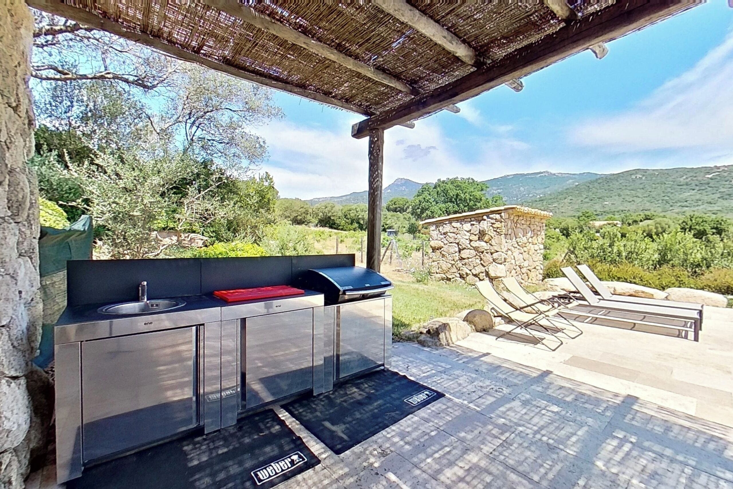 An outdoor kitchen with a sink and a grill, surrounded by a stone wall and a pergola.