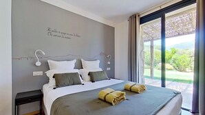 Une chambre à coucher avec un lit, une table de chevet, une lampe fixée au mur et une vue sur la verdure par la fenêtre.