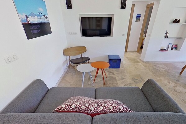 Una sala de estar con un sofá gris, una mesita con dos sillas, un televisor montado en la pared y una estantería.