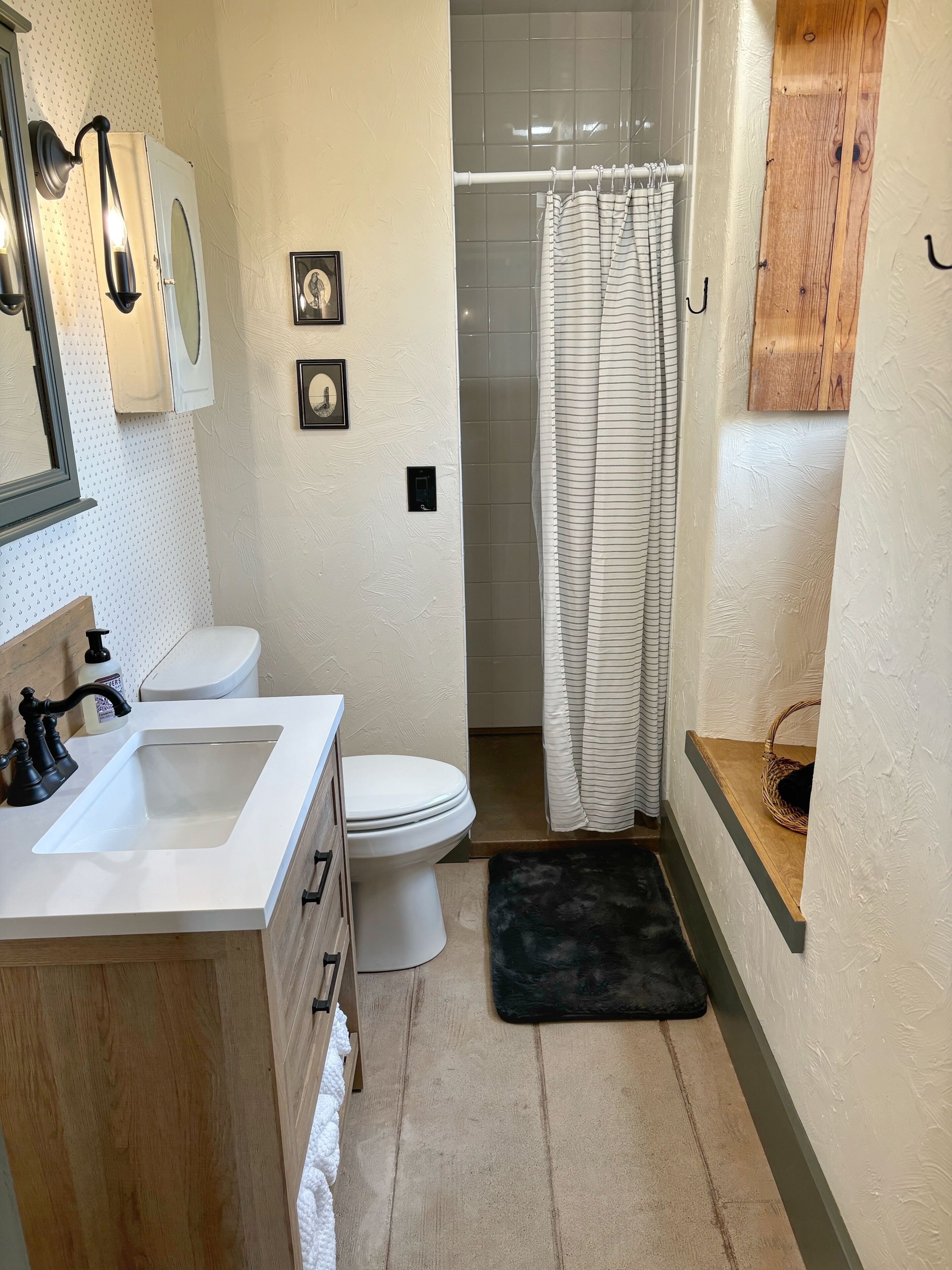 A bathroom with a white sink, toilet, and shower curtain. There are framed pictures on the wall and a wooden shelf with a basket.