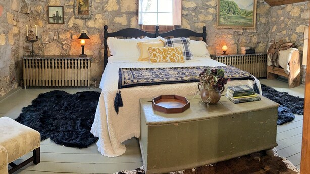 Una camera da letto rustica con parete in pietra, soffitto in legno, un letto con molti cuscini, un comodino con libri e una finestra vista.