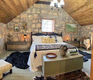 Un dormitorio rústico con pared de piedra, techo de madera, cama con muchos cojines, mesita de noche con libros y ventana con vistas.