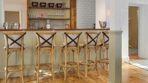 Eine Bar mit Holzhockern, einer weißen Deckenleuchte und einem Regal mit schwarzen Eimern und Gläsern.