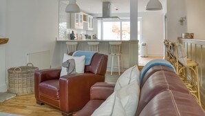 Ein Wohnzimmer mit einer braunen Ledersofa, einem weißen Sofa und einem Korbwannenstuhl. Es gibt eine Küchenzeile mit weißen Schränken und einem Fenster.