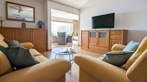 A spacious living room with a sofa, a coffee table, and a television. There is a balcony with a view of buildings.