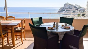 A balcony with a table set for two, overlooking a coastal view with a prominent hill in the distance.