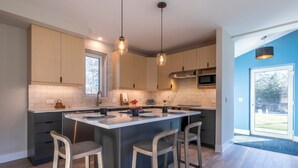 Una cocina moderna con una isla central, taburetes de bar, iluminación colgante y vistas al exterior.
