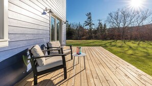 Una terraza de madera con un sofá y una mesa, una casa de revestimiento blanco y un cielo despejado.