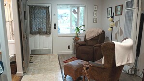 A living room with a brown armchair, a small ottoman, a potted plant, and a window with blinds.