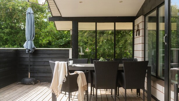 A covered outdoor dining area with a table and chairs, a wooden deck, and a view of greenery through large windows.