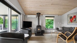 A modern living room with a fireplace, comfortable seating, a flat-screen TV, and large windows offering a view of greenery.