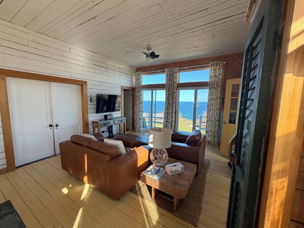 A room with a wooden ceiling, a television, a sofa, a coffee table, and a view of the sea through large windows. - Former one-room schoolhouse with a 180° ocean view • Near Forillon (Gaspé)