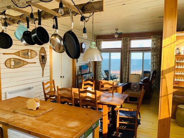 A wooden dining area with a table and chairs, a kitchen with hanging pots and pans, and a view of the sea through large windows. - Breathtaking ocean views, peace, and relaxation (Gaspé)