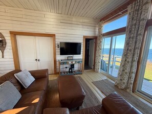 A cozy living room with a brown leather sofa, a flat-screen TV on a wooden stand, and a large window with curtains. - Former one-room schoolhouse with a 180° ocean view • Near Forillon (Gaspé)