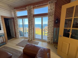 A room with a wooden ceiling, a brown leather sofa, a yellow door, and a view of the sea through large windows. - Former one-room schoolhouse with a 180° ocean view • Near Forillon (Gaspé)