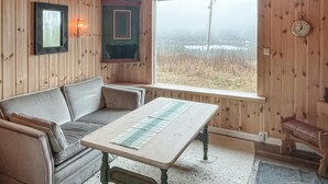 Een knus ingerichte kamer met houten details, een bank, een tafel en uitzicht op een veld door een groot raam.