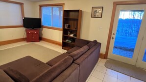 A living room with a sectional sofa, a television on a stand, a bookshelf, and a door leading outside. - Cozy lakefront cabin on Big Beaver Lake (Big Lake)