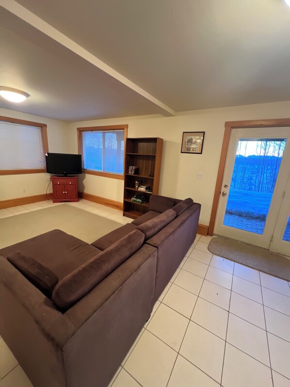 A living room with a sectional sofa, a television on a stand, a bookshelf, and a door leading outside. - Cozy lakefront cabin on Big Beaver Lake (Big Lake)