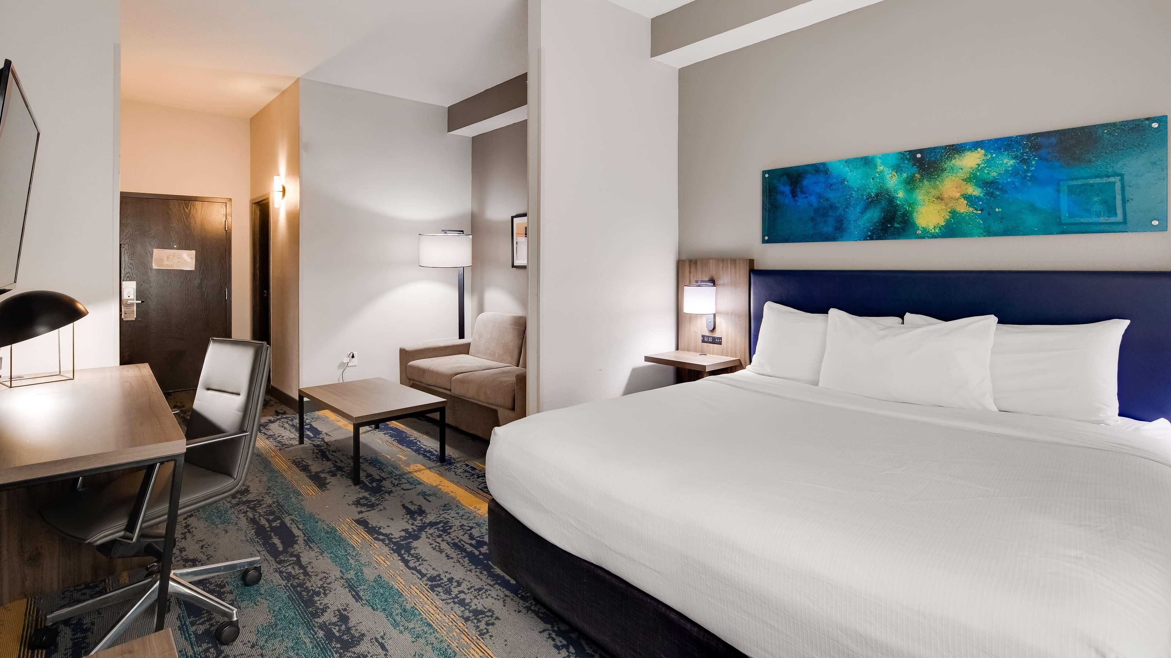 A hotel room with a bed, a desk, a chair, a small table, a lamp, and a painting on the wall.