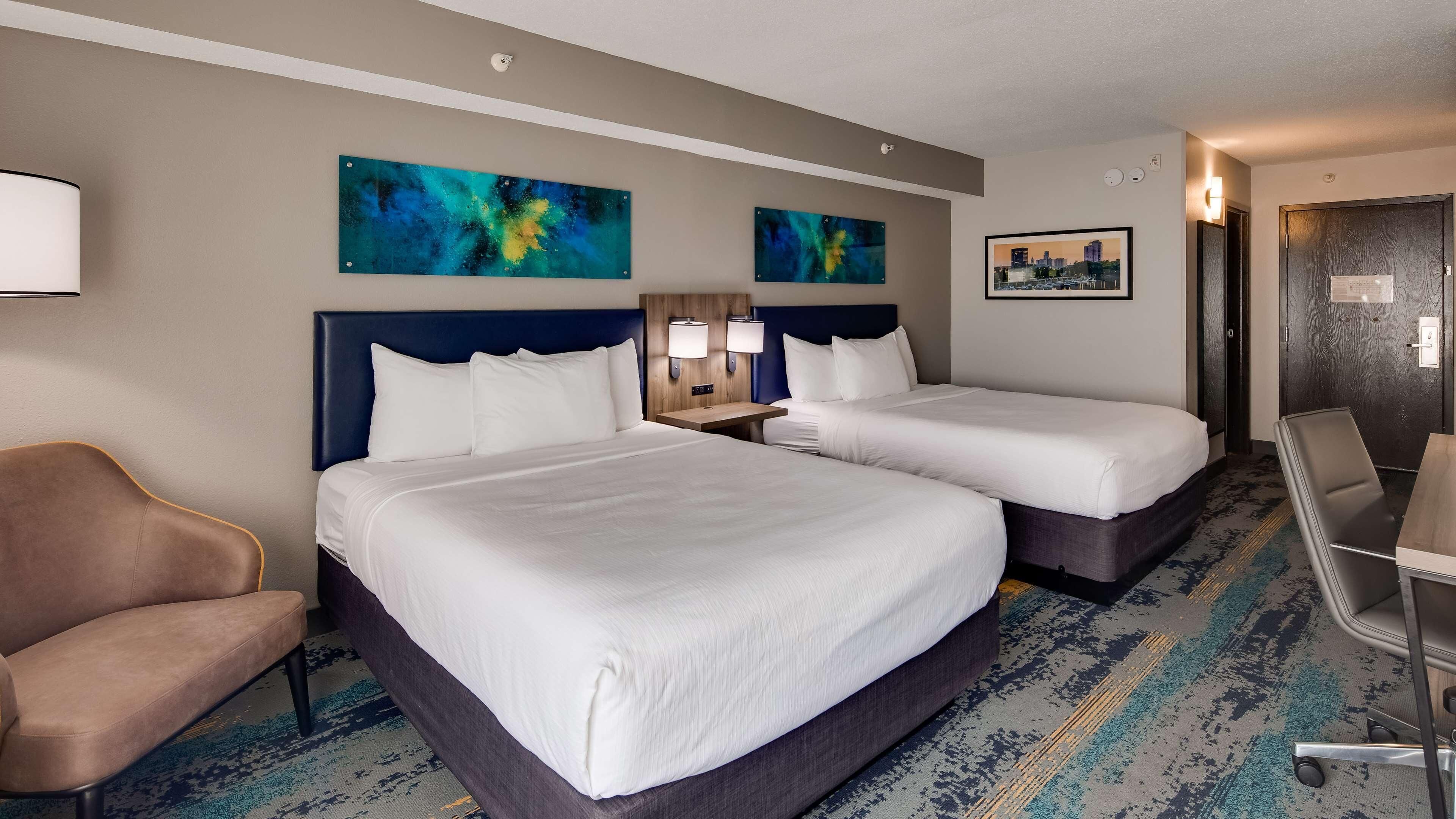 A hotel room with two beds, a desk, a chair, and artwork on the walls.