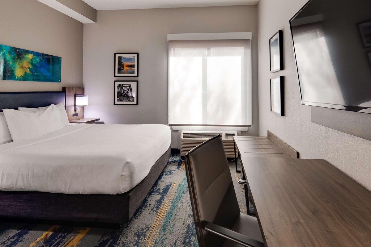 A hotel room with a bed, a desk, a chair, a TV, a window, and artwork on the walls.