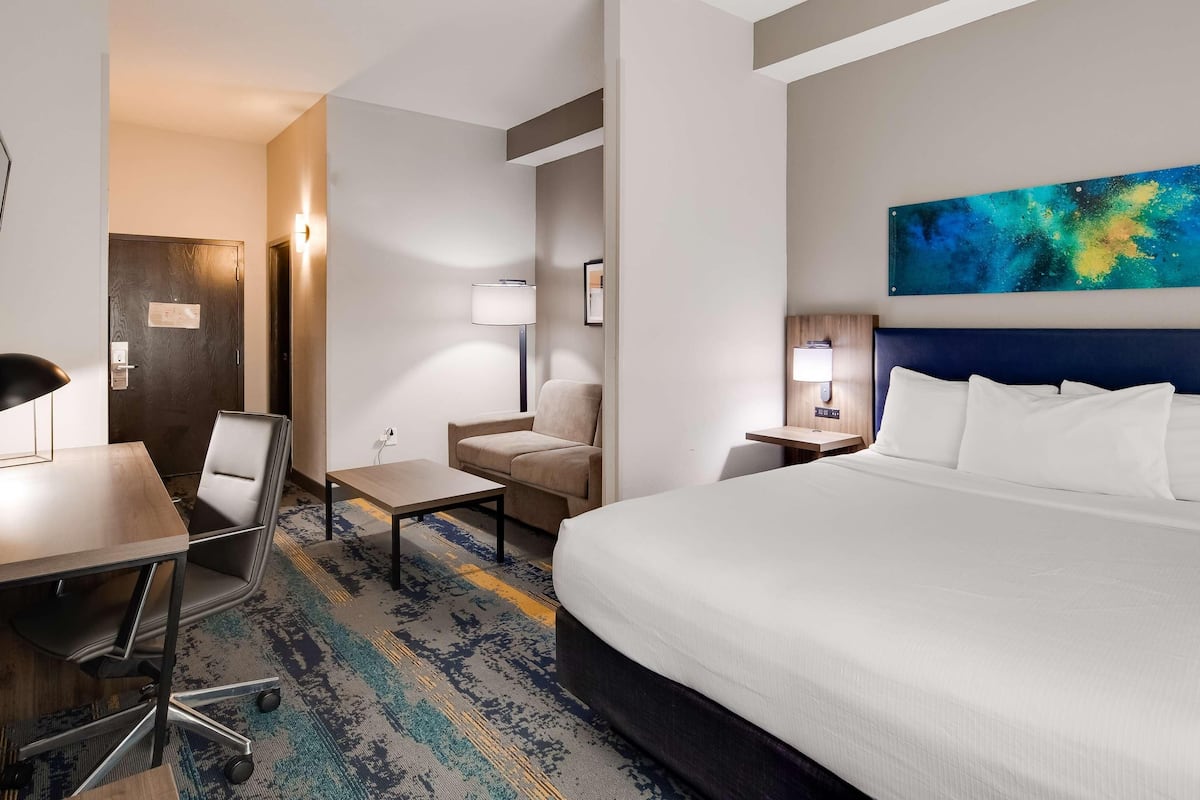 A hotel room with a bed, a desk, a chair, a small table, a lamp, and a painting on the wall.