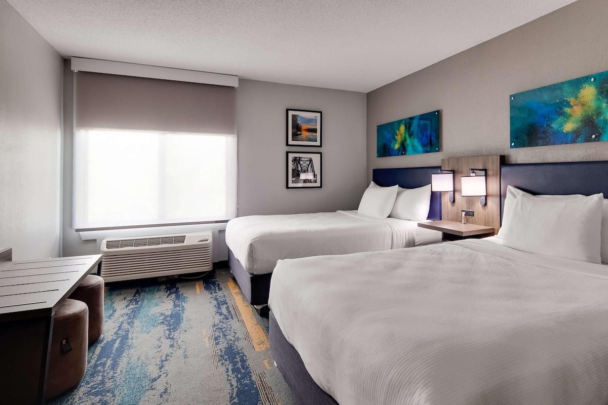 A hotel room with two beds, a desk, a chair, a window with blinds, a wall-mounted air conditioner, and two paintings.