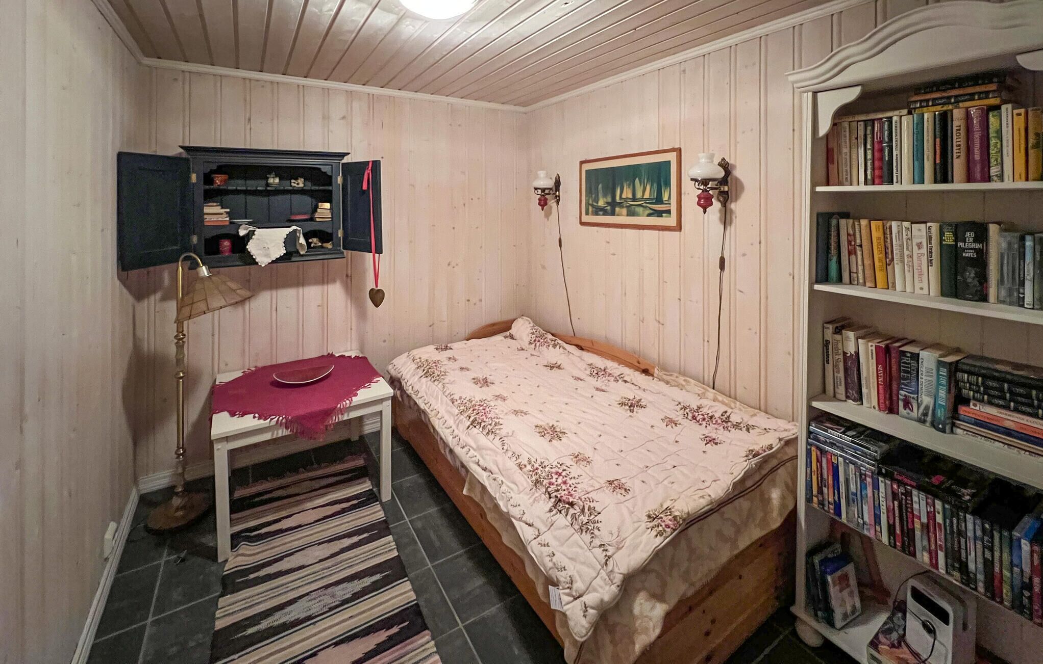 Ett litet sovrum med en enkelsäng, en bokhylla, ett skrivbord med en lampa och en mönstrad matta.