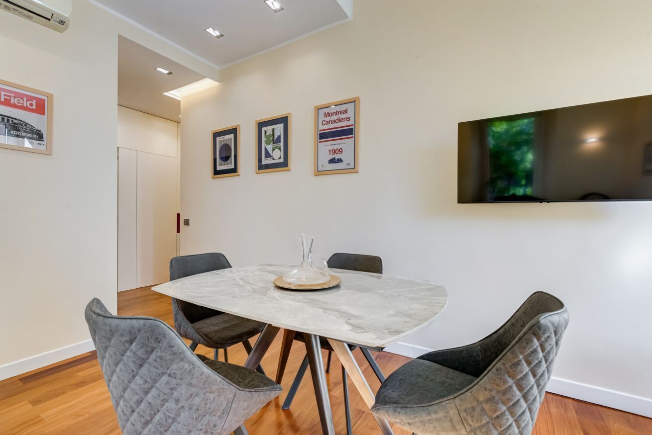 A dining area with a marble table, four cushioned chairs, a mounted flat-screen TV, and framed pictures on the wall.
