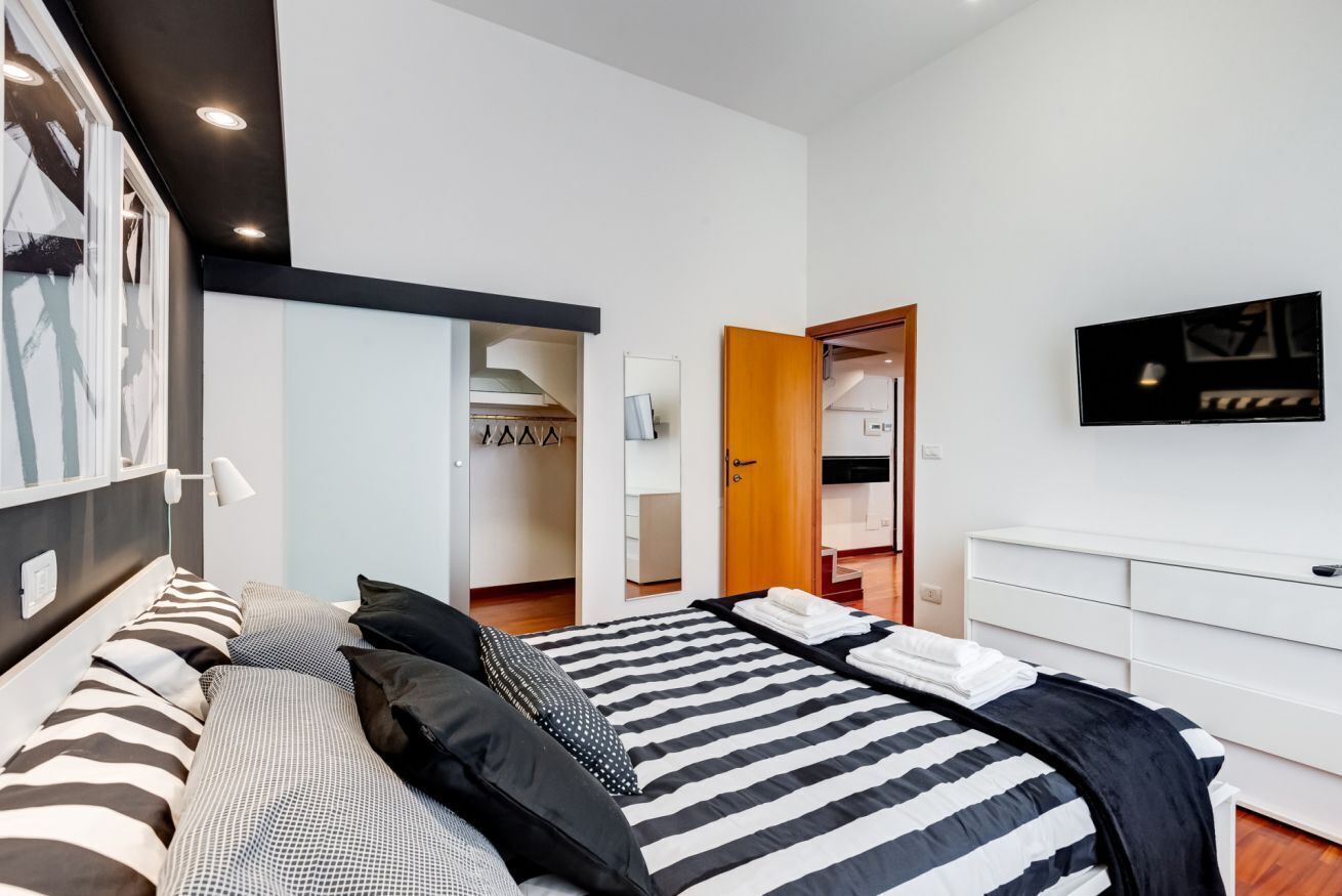 A modern bedroom with a large bed, a wardrobe, a mirror, and a television on the wall.
