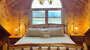 A wooden cabin bedroom with a bed, a chandelier, and a window with a view. - Secluded lakeside retreat with lake view and hot tub. Close to Mammoth Cave.  (Cub Run)
