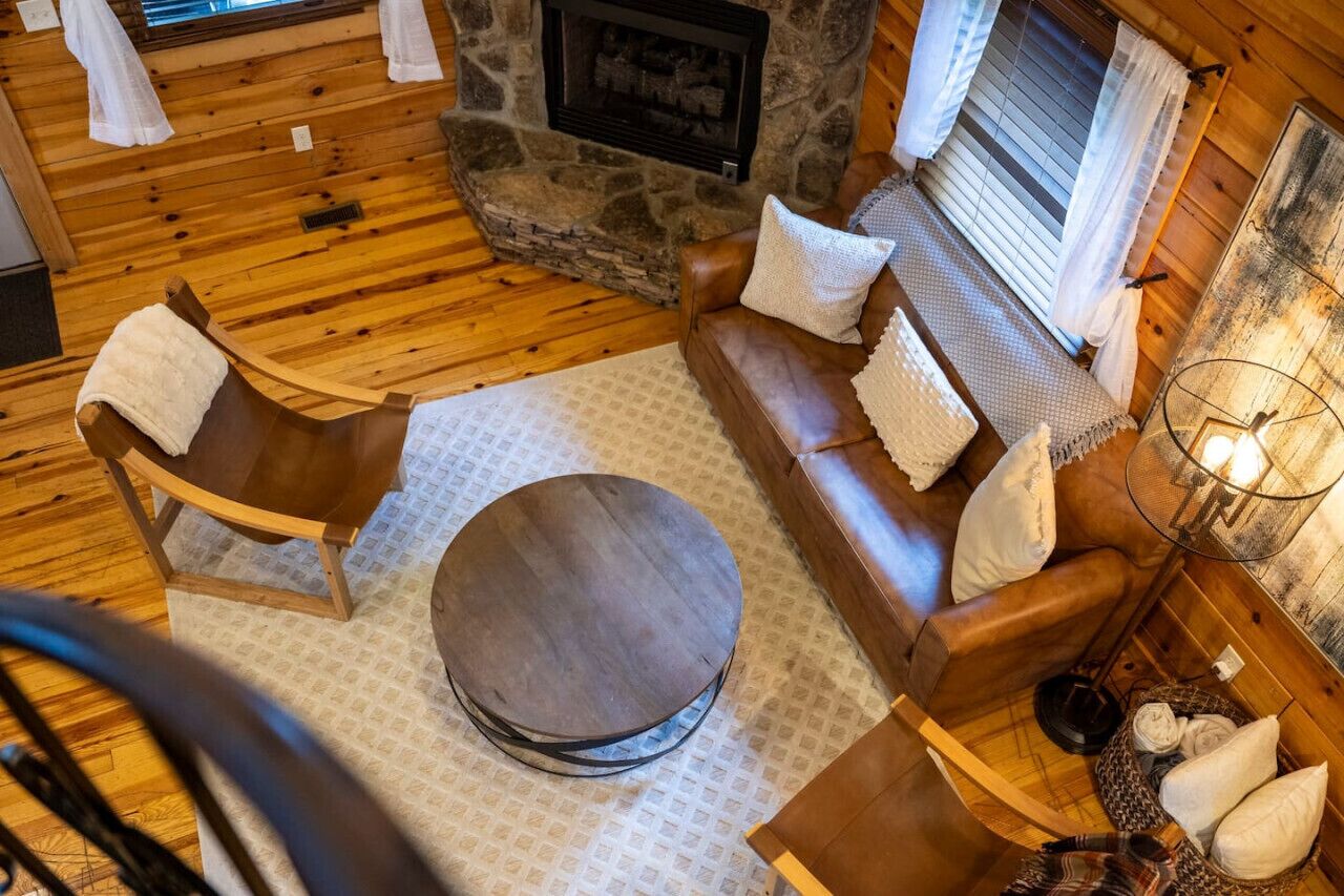 A cozy cabin interior with a stone fireplace, wooden flooring, a round ottoman, leather sofa, and a lamp.