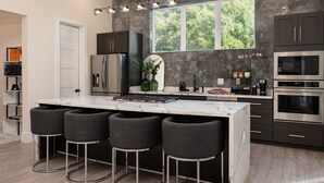 A modern kitchen with a marble island, bar stools, built-in appliances, and a large window with a view of trees. - Designer Luxury for 10•Rooftop Deck•Rice Military (HOUSTON)
