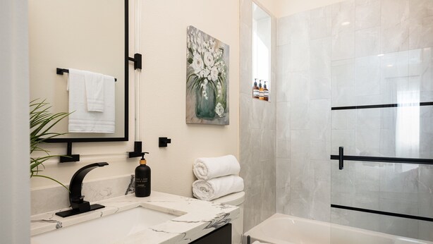 Ein modernes Badezimmer mit Marmorwaschbecken, einem schwarzen Wasserhahn, einem weißen Handtuch, einem Blumenbild und einer Duschkabine aus Glas.
