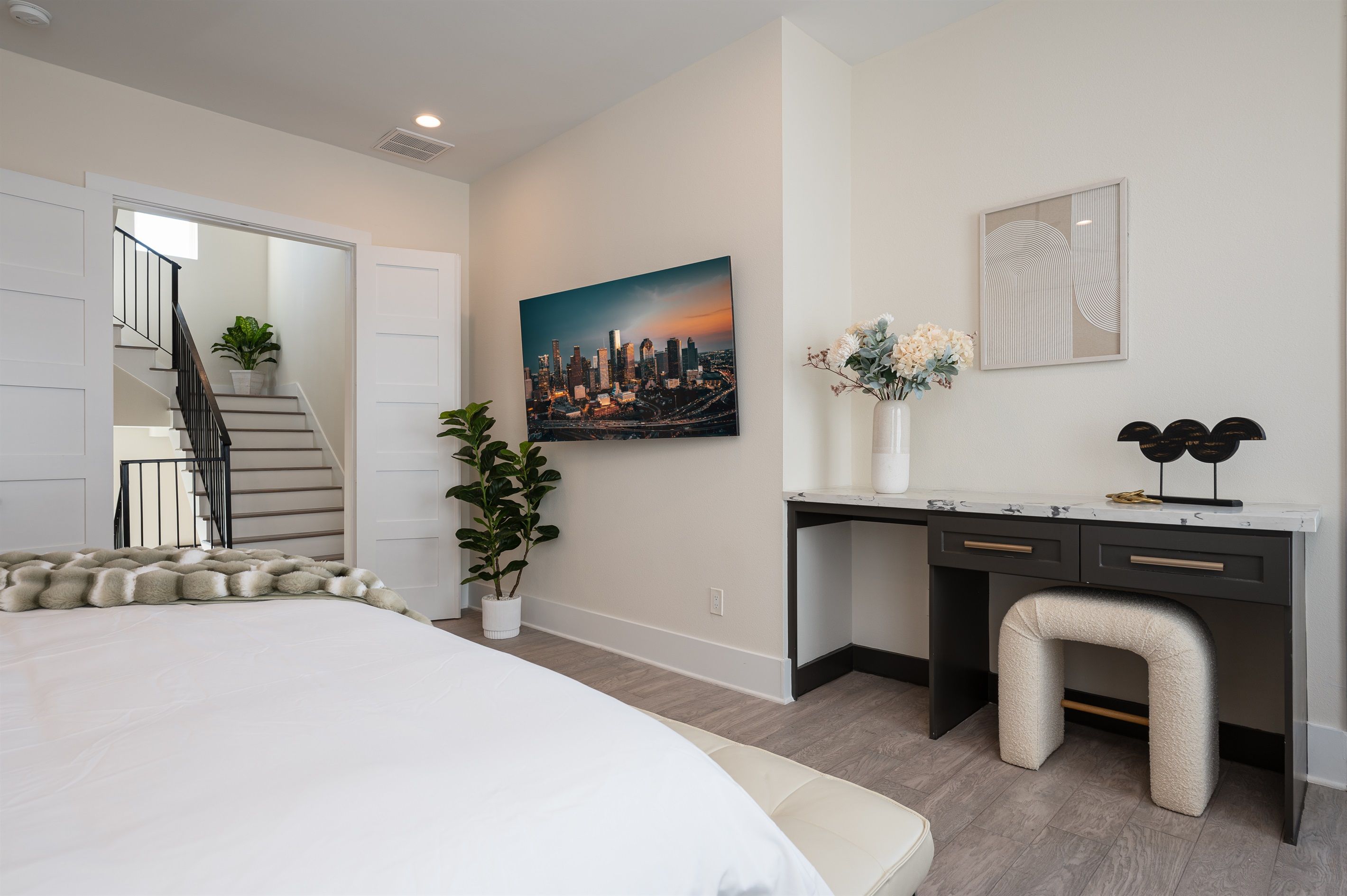 A bedroom with a bed, a desk, a chair, a potted plant, and a painting of a cityscape.