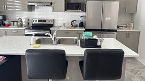 Een moderne keuken met grijze kastjes, roestvrijstalen apparatuur, een centraal eiland met twee zwarte stoelen en een betegelde achterwand.