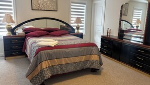 Een slaapkamer met een groot bed, twee nachtkastjes, een dressoir, een spiegel en een raam met jaloezieën.