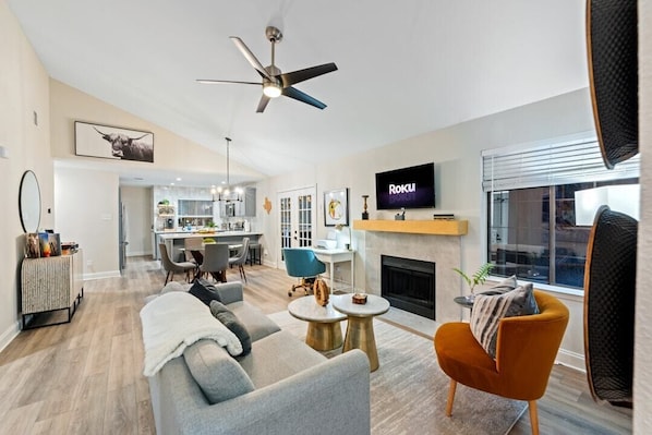 A modern living room with a sofa, armchairs, and a fireplace. There is a TV mounted on the wall and a ceiling fan.