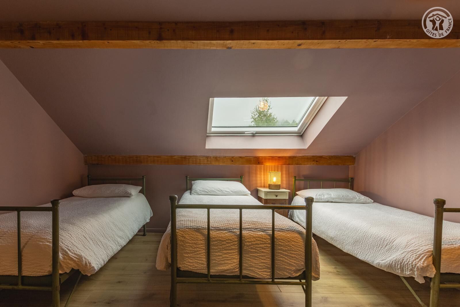 A cozy attic bedroom with three wooden beds, a skylight, and a nightstand with a lamp.