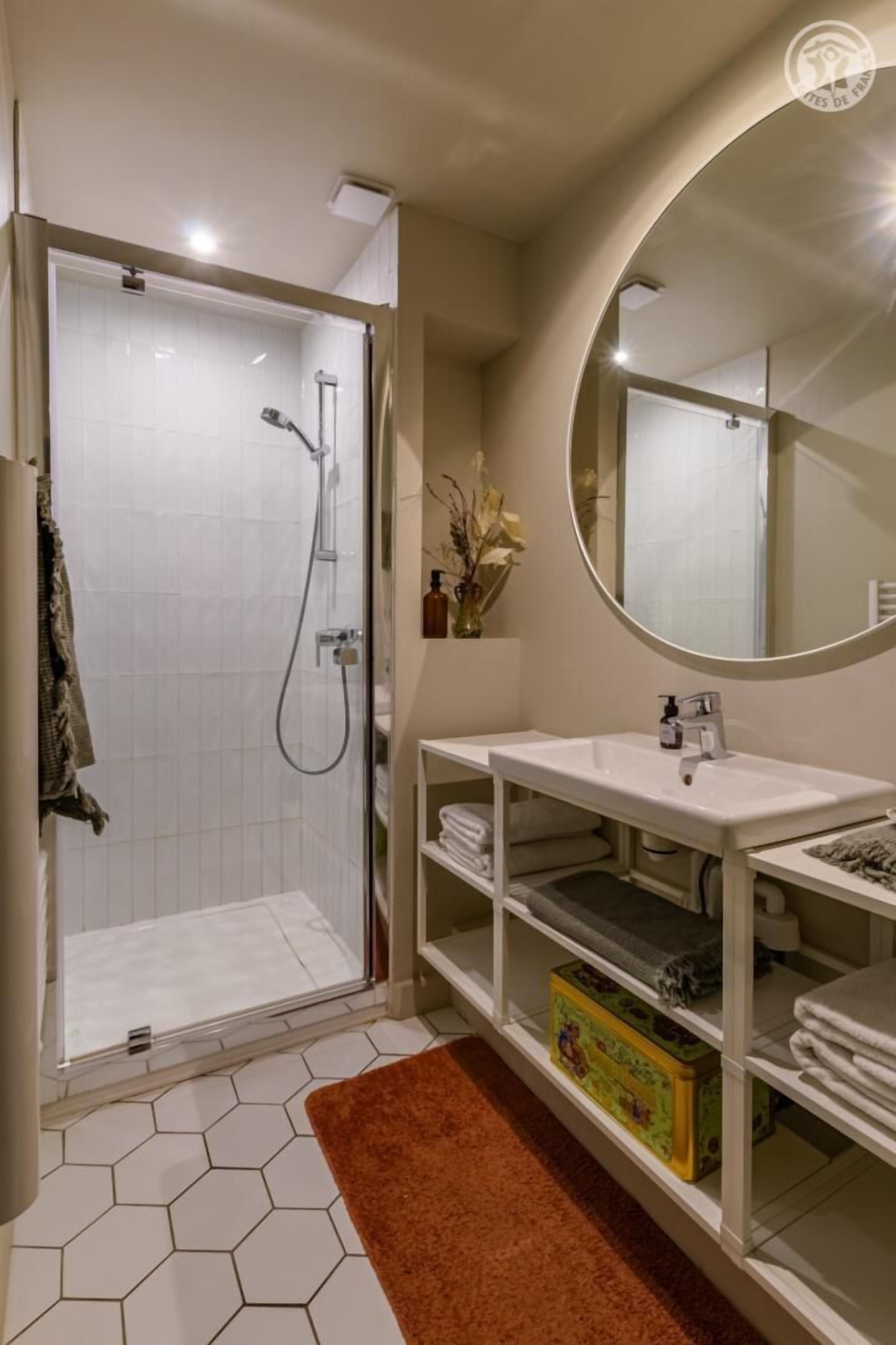 A modern bathroom with a glass-enclosed shower, a white sink, a round mirror, and a hexagonal tile floor.