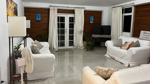 A living room with two white sofas, a TV, and a fan. There are potted plants and a painting on the wall. - Modern Eco lodge with A/C in bedrooms and 40ft patio for relaxation and fun. (St. Sylvans Village)