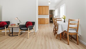Una cocina moderna con armarios de madera, un comedor con una mesa redonda y sillas, y una zona de estar acogedora con una lámpara y cojines.