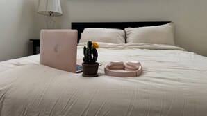 Una cama bien hecha con una laptop, un pequeño cactus y un par de pantuflas sobre la mesita de noche.