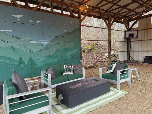 An outdoor seating area with green cushioned sofas, a black fire pit, and a mural of a mountainous landscape. - Creekside Mountain Cabin Getaway - 6 Acres, Mountain Views, Hot Tub (Cosby)