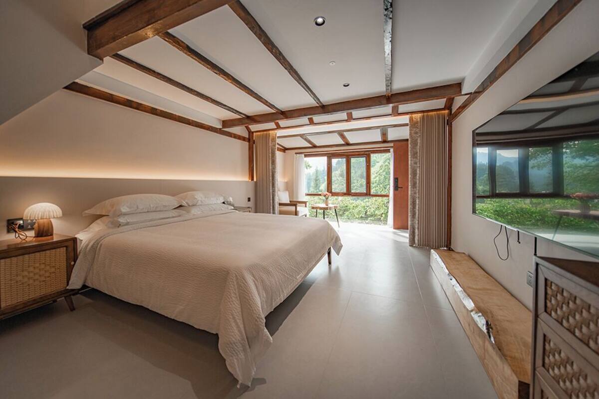 A bedroom with a large bed, wooden beams, a bedside table with a lamp, and a view of greenery through a window.