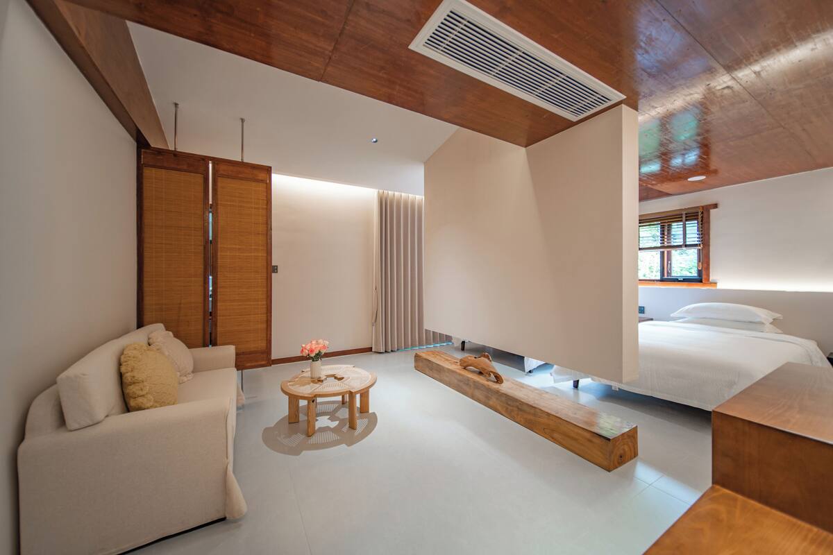 A modern hotel room with a wooden ceiling, a sofa, a small round table with a vase of flowers, a wooden bench, and a bed with a window view.