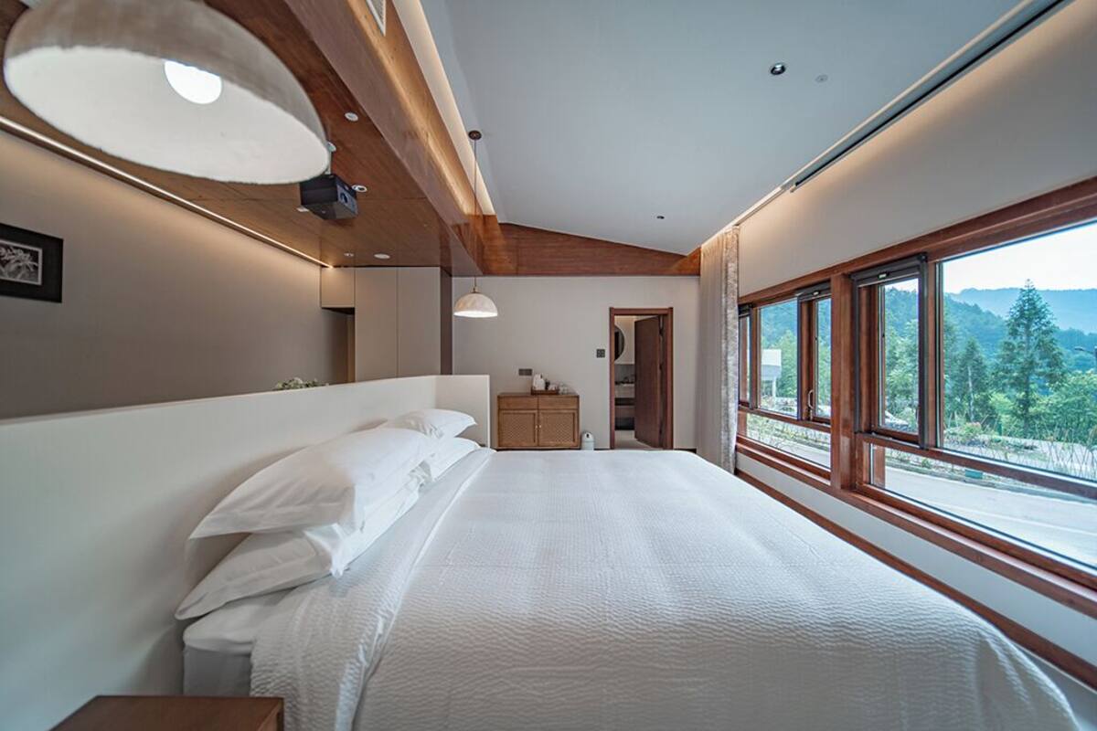 A bedroom with a large bed, wooden ceiling beams, a wooden dresser, and a view of the outdoors through large windows.