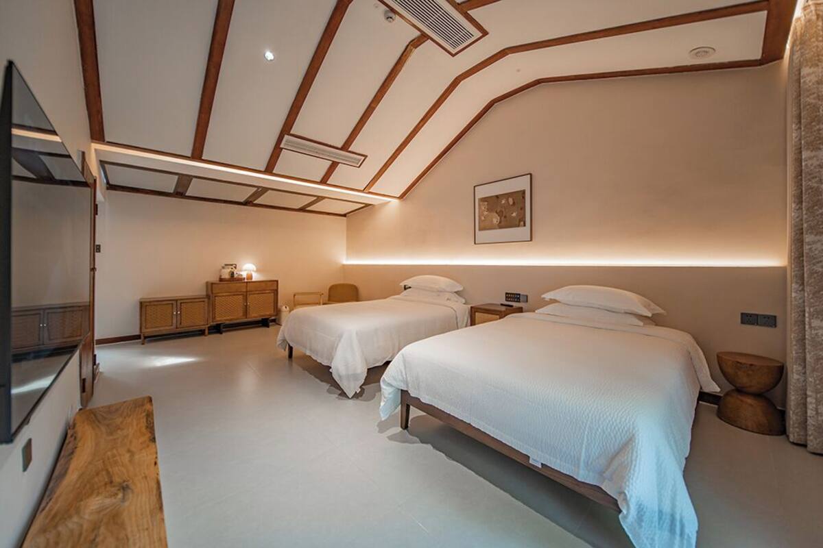 A modern bedroom with two beds, a wooden bench, a TV, and a ceiling with exposed beams and recessed lighting.