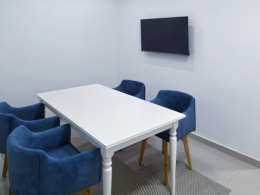 Un salón de conferencias con una mesa blanca, sillas azules, un televisor de pantalla plana en la pared y una alfombra estampada.